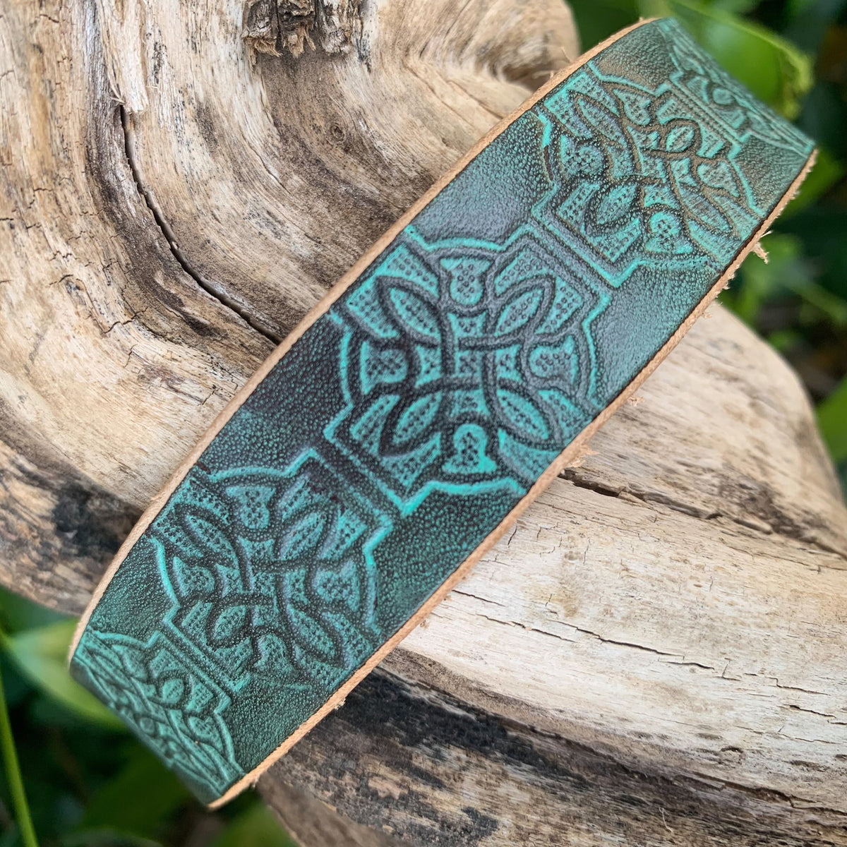 A wide leather dog collar with embossed Celtic knot patterns in a dark green and turquoise color combination, displayed on a rustic log with green foliage in the background.