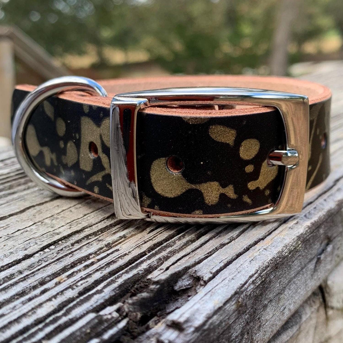 A leather dog collar with a black and tan abstract pattern, silver-toned metal buckle, and a brown leather underside, resting on a weathered wooden surface with a blurred outdoor background.