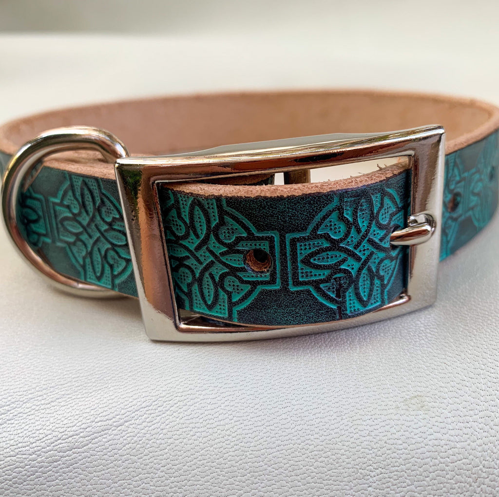 A close-up of a leather dog collar with a turquoise and black Celtic knot pattern, a silver-tone buckle, and a tan leather underside, resting on a white textured surface.