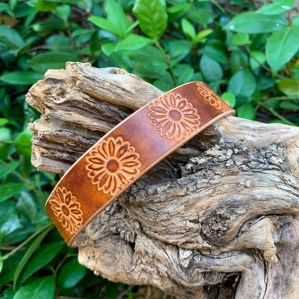 A wide brown leather dog collar with an embossed flower pattern, resting on a piece of weathered driftwood with green foliage in the background.