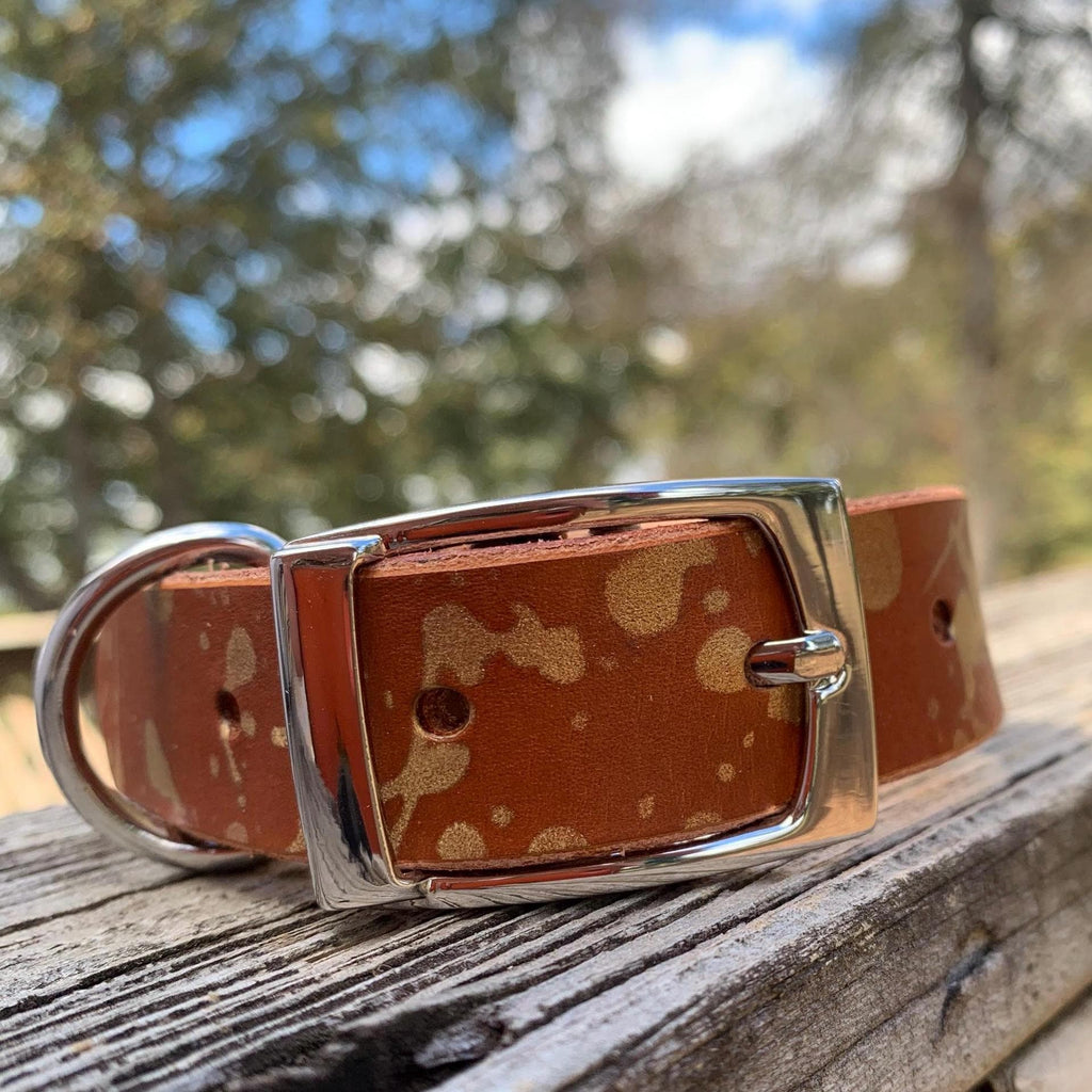 A brown leather dog collar with a tan and gold drip pattern and a silver-tone buckle, resting on a weathered wooden surface with a blurred outdoor background.