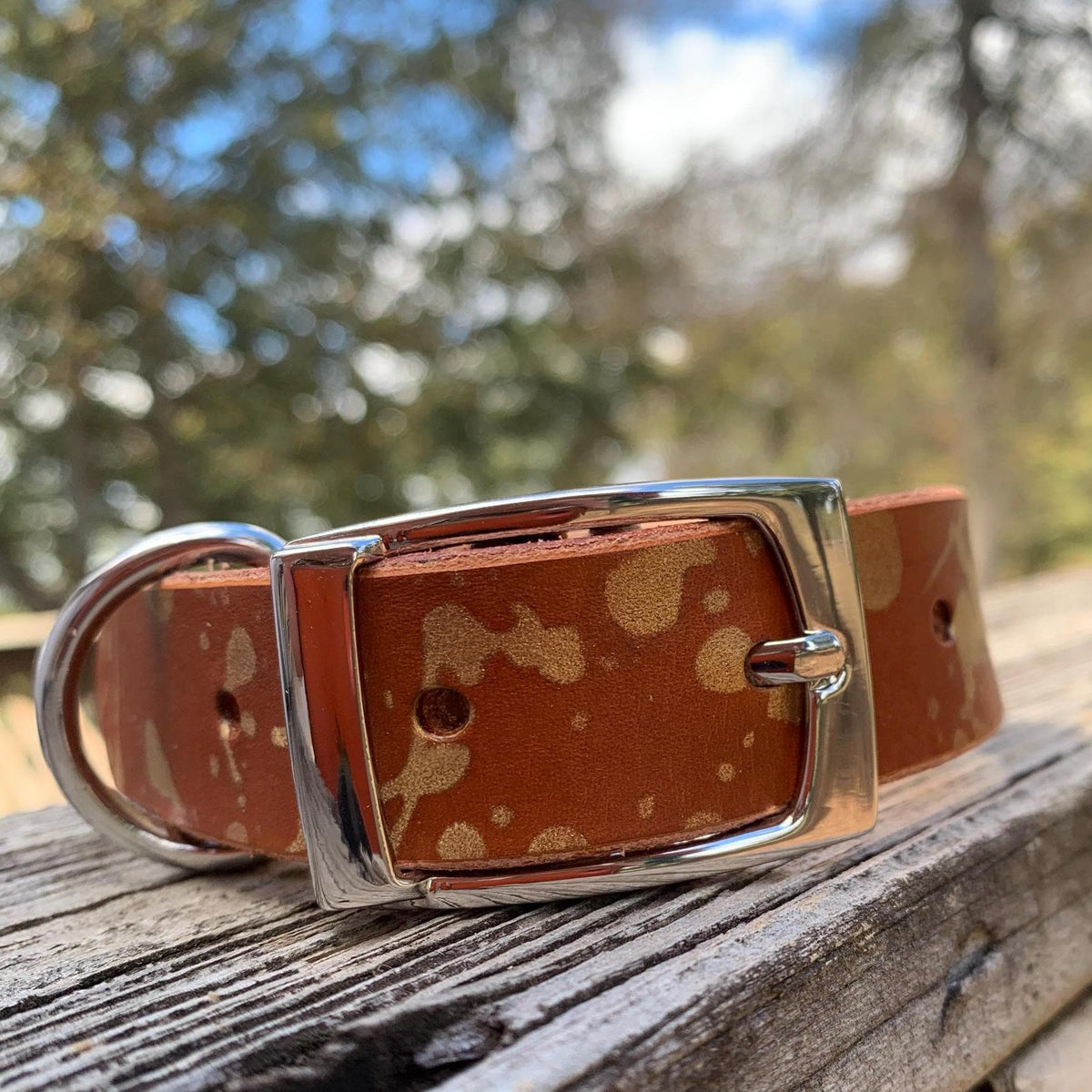 A brown leather dog collar with a tan and gold drip pattern and a silver-tone buckle, resting on a weathered wooden surface with a blurred outdoor background.