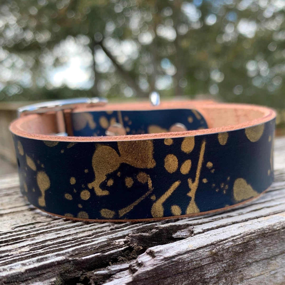 A dark navy blue leather dog collar with a gold abstract paint splatter pattern, resting on a weathered wooden surface with a blurred outdoor background.
