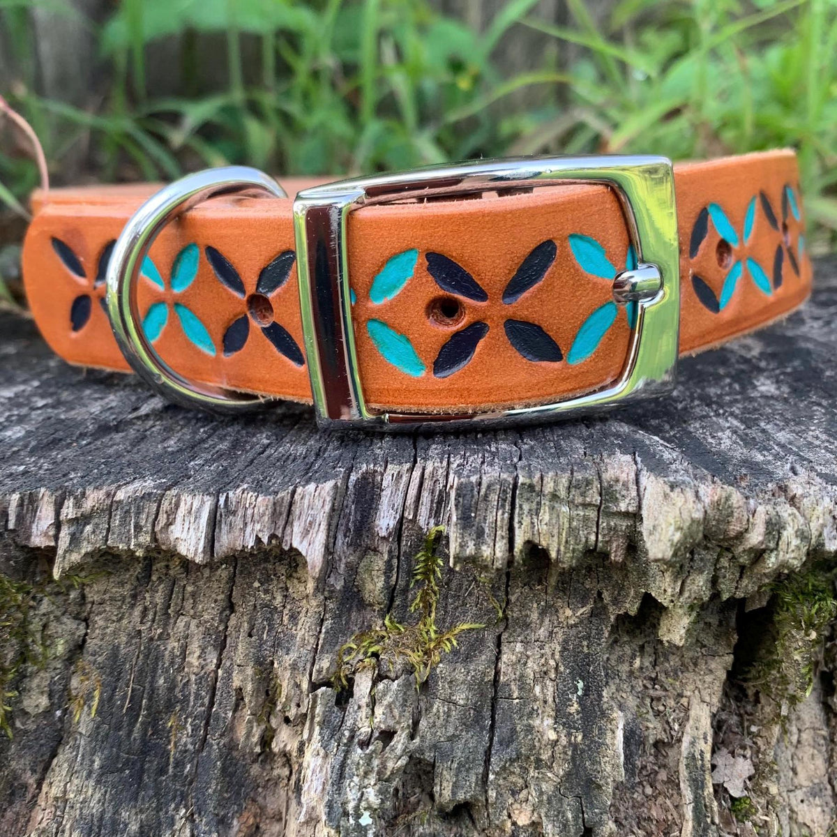A medium brown leather dog collar with turquoise and navy blue geometric pattern, resting on a weathered tree stump with green foliage in the background.