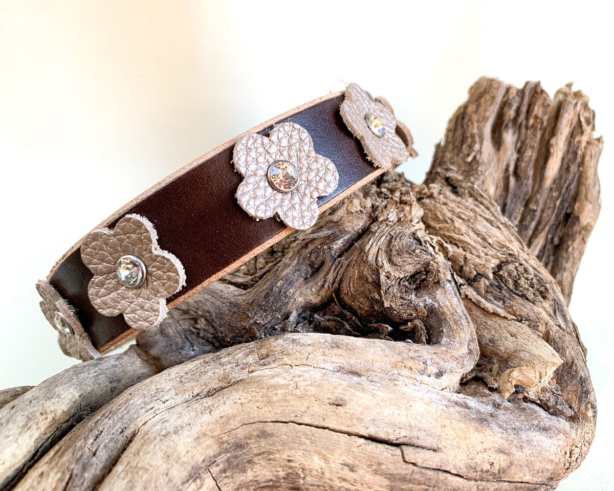 A dark brown leather dog collar textured beige flower appliqués and small rhinestone centers, displayed on a piece of weathered driftwood.