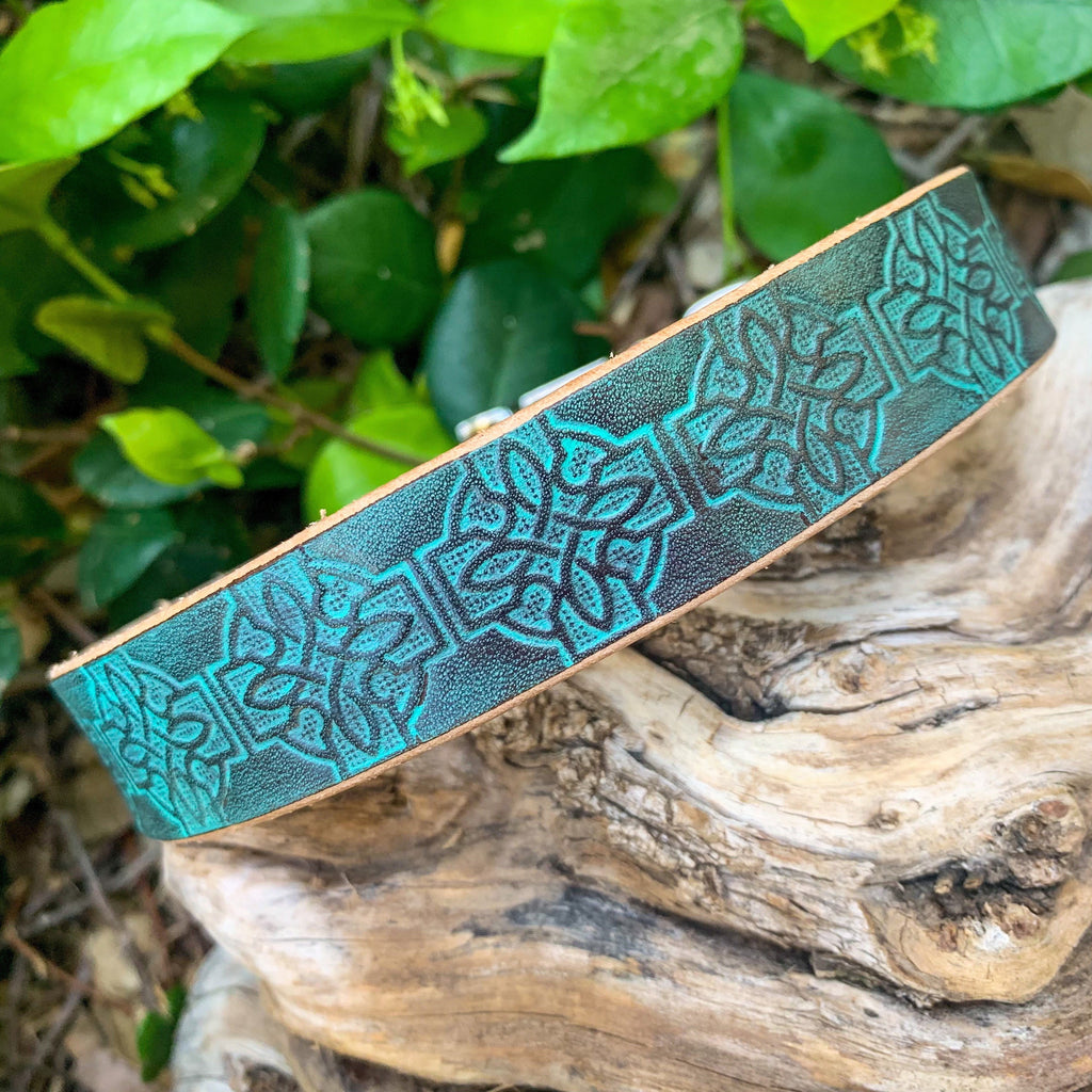 A turquoise leather dog collar with black embossed Celtic knot patterns, resting on a piece of weathered wood with green foliage in the background.