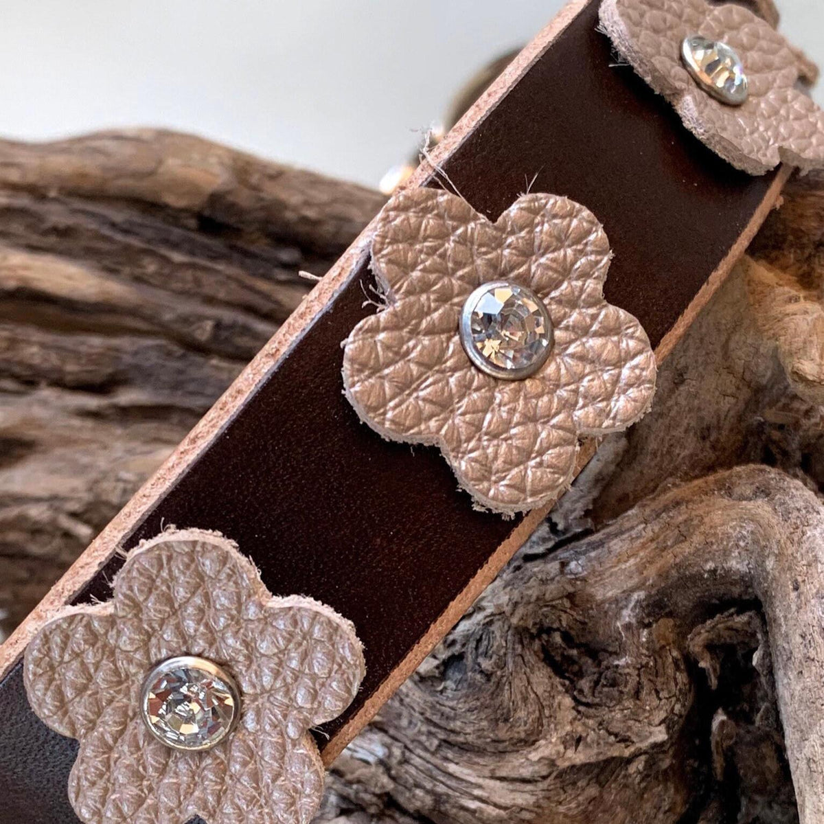 A close-up of a dark brown leather strap with three textured beige flower-shaped leather embellishments, each set with a clear rhinestone, resting on a piece of driftwood.