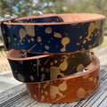 Three leather dog collars stacked on a weathered wooden surface, featuring a navy blue top collar, black center collar and brown bottom collar, all decorated with a metallic gold paint splatter pattern.