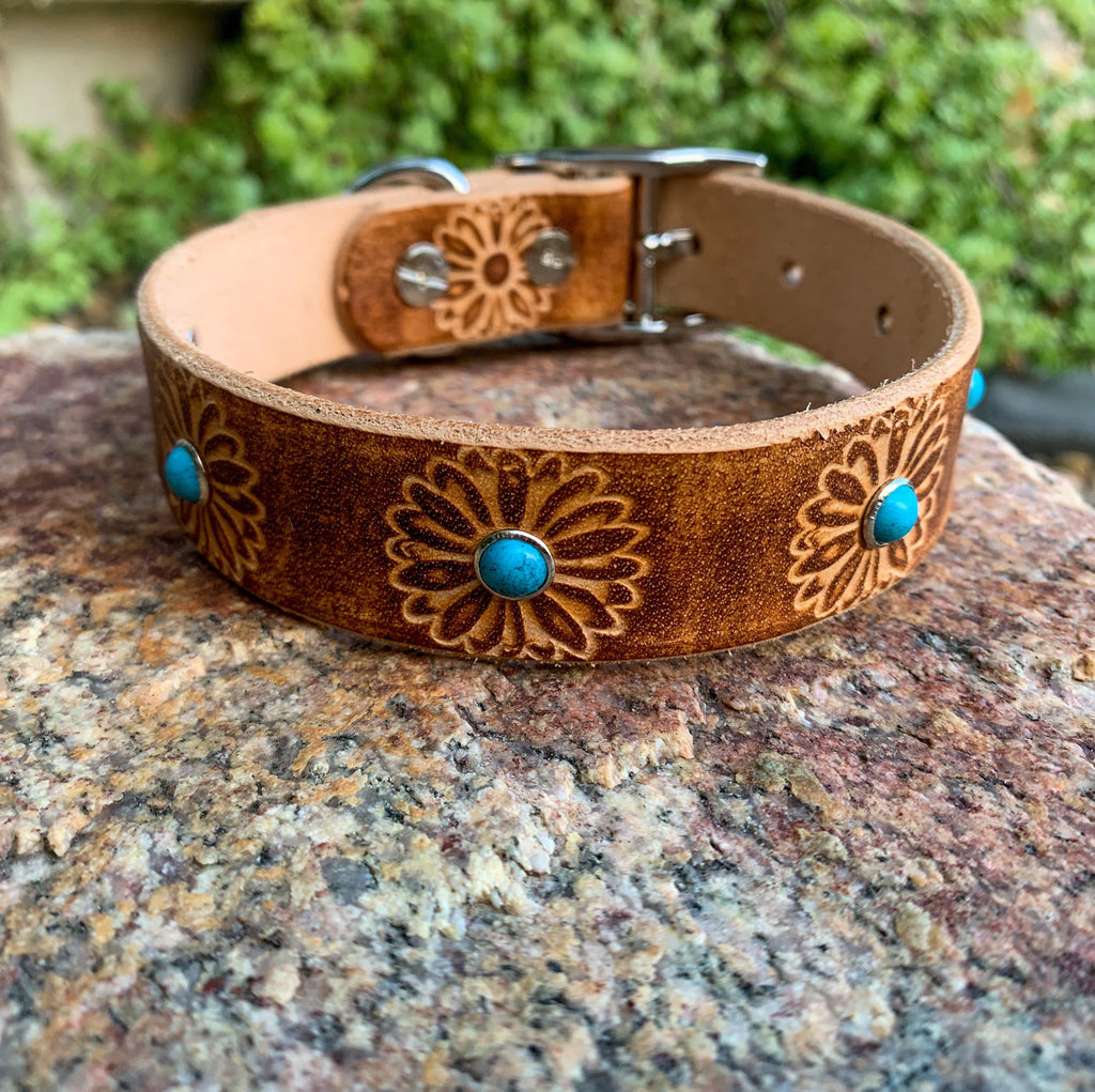 A brown leather dog collar with floral engravings and turquoise stone accents, resting on a textured rock with a blurred green garden background.