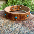 A brown leather dog collar with floral engravings and turquoise stone accents, resting on a textured rock with a blurred green garden background.