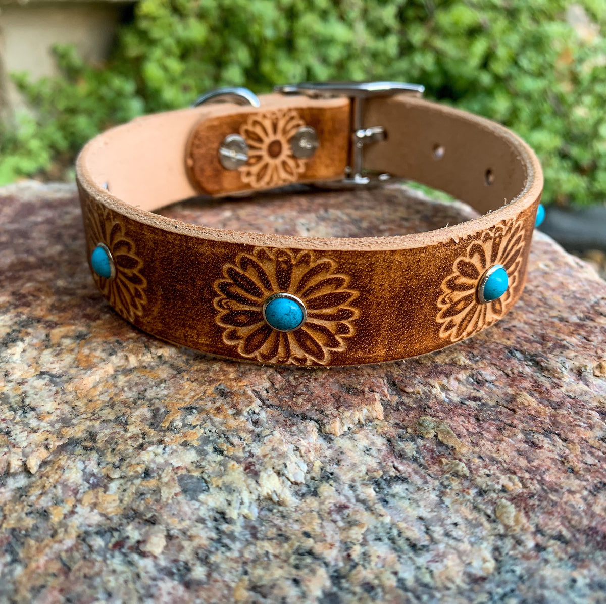 A brown leather dog collar with floral engravings and turquoise stone accents, resting on a textured rock with a blurred green garden background.