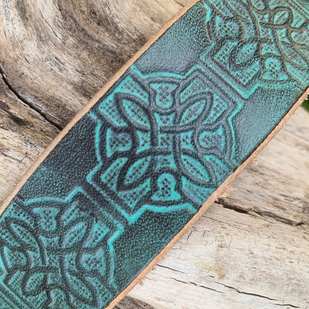 A close-up of a leather strap or belt featuring a textured turquoise and black Celtic knot pattern, laid diagonally on a rustic weathered wood surface.