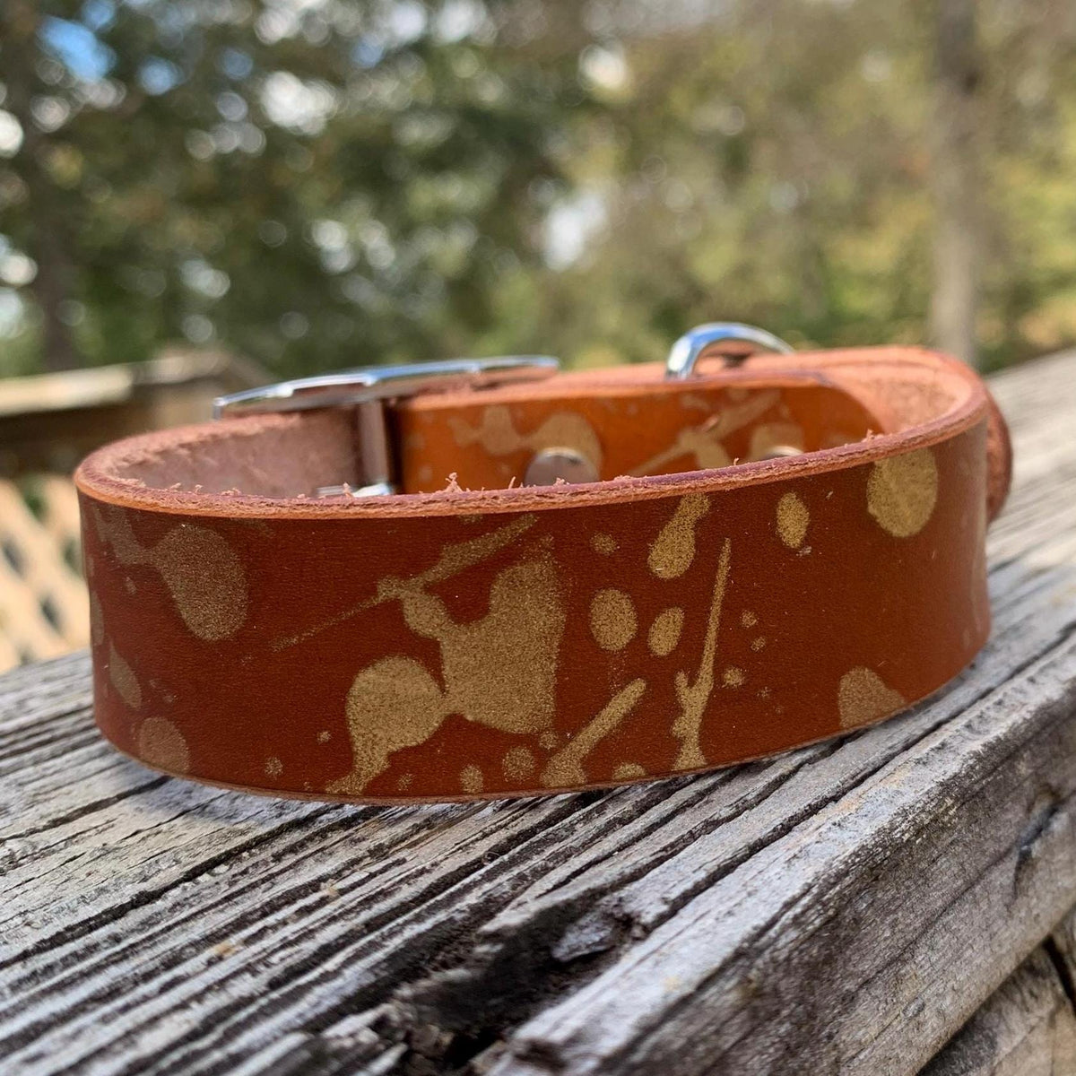 A brown leather dog collar with a gold paint-splatter pattern and a silver metal buckle, resting on a weathered wooden deck with a blurred outdoor background.