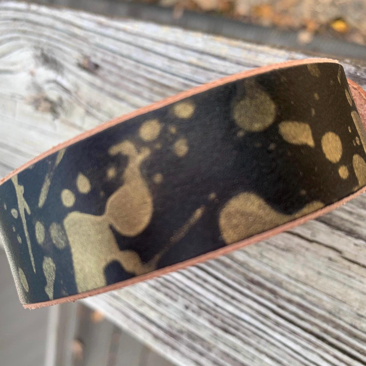 A close-up of a wide leather dog collar with a black and gold paint-splatter pattern, resting on a weathered gray wooden surface.