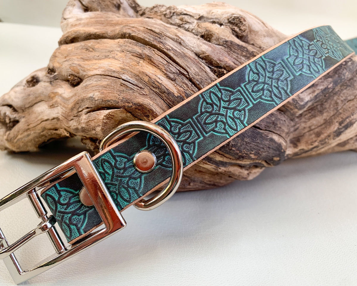 A close-up of a leather dog collar with a turquoise and brown Celtic knot pattern, resting on a piece of natural driftwood over a white surface.