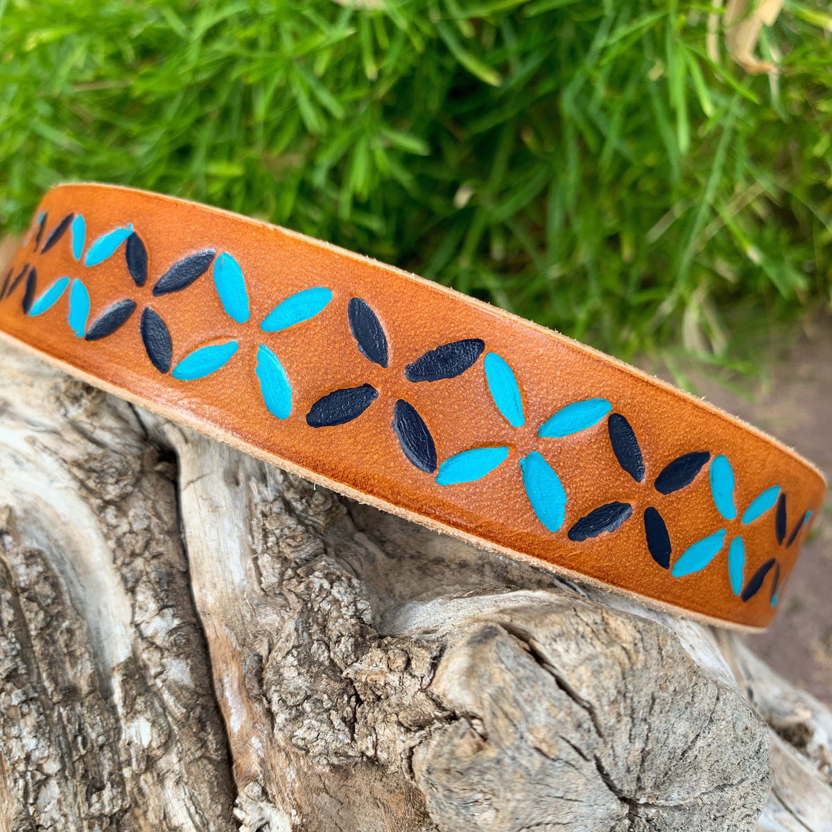 A tan leather dog collar with a hand-painted pattern of turquoise and black geometric shapes, resting on a piece of weathered wood with green foliage in the background.