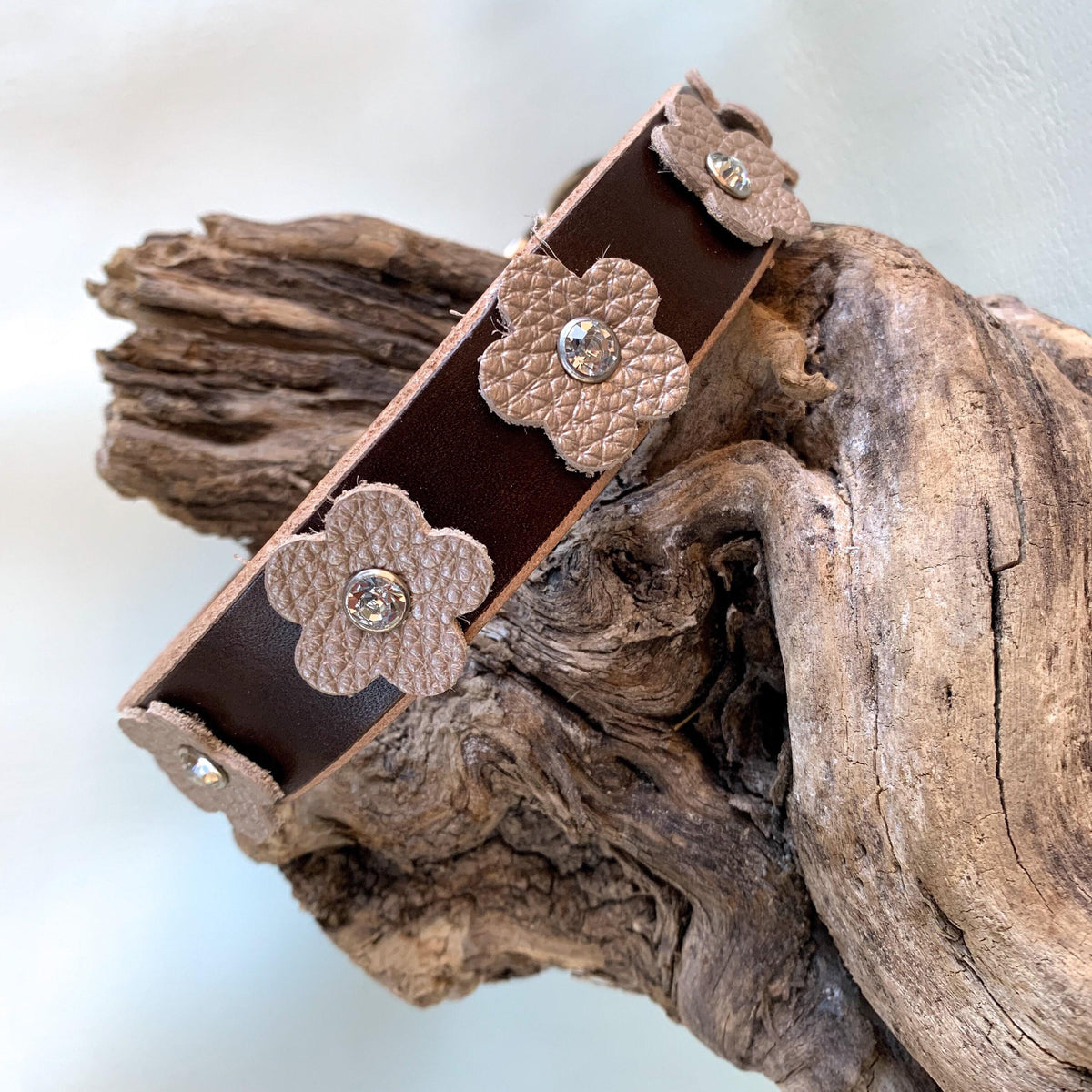 A dark brown leather dog collar with textured beige flower appliqués and small rhinestone centers, displayed on a piece of weathered driftwood.