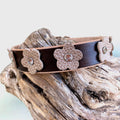 A dark brown leather dog collar textured beige flower appliqués and small rhinestone centers, displayed on a piece of weathered driftwood.