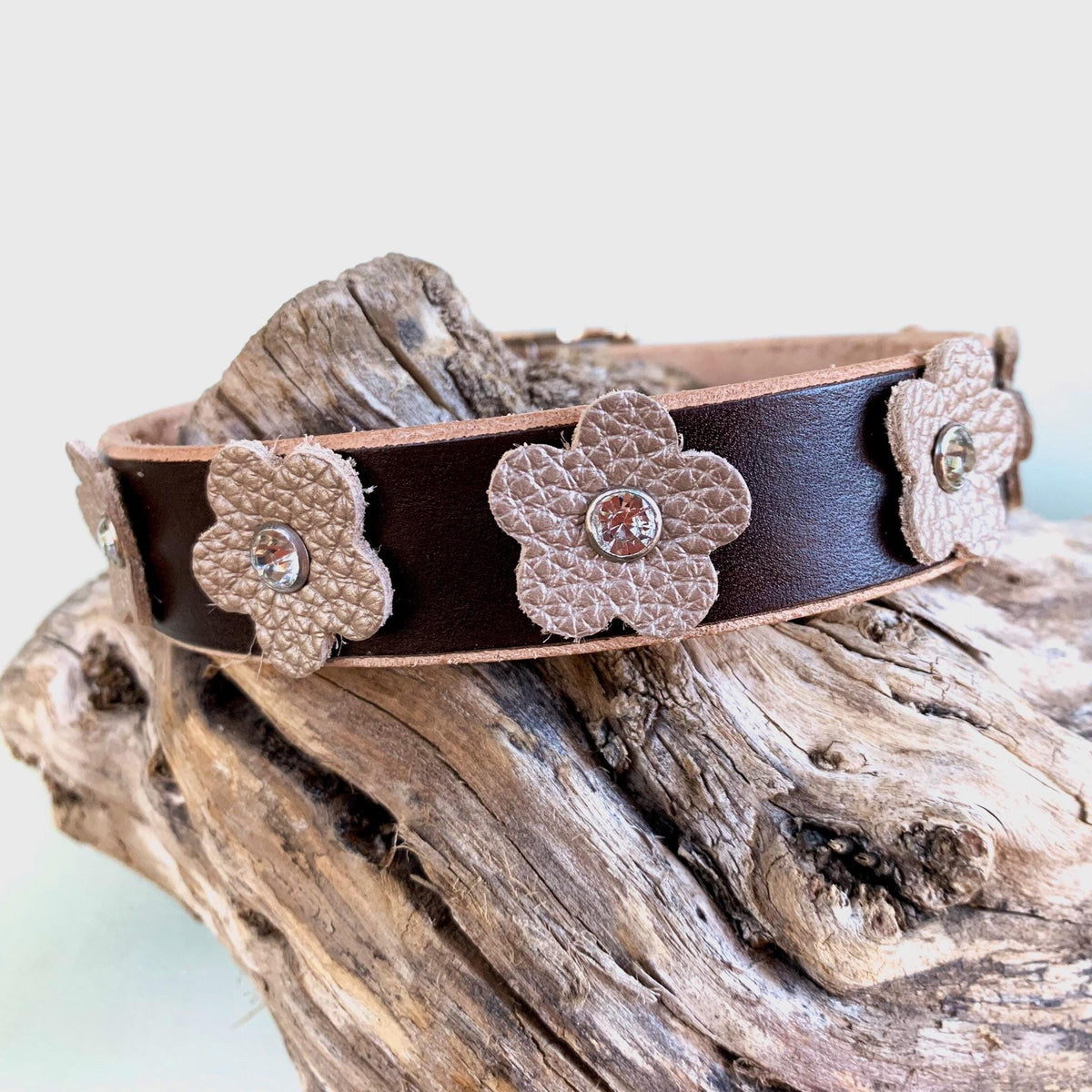 A dark brown leather dog collar textured beige flower appliqués and small rhinestone centers, displayed on a piece of weathered driftwood.