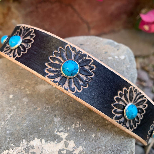 A black leather dog collar with hand-tooled floral designs and turquoise stone accents, displayed on a gray stone with a blurred outdoor background.
