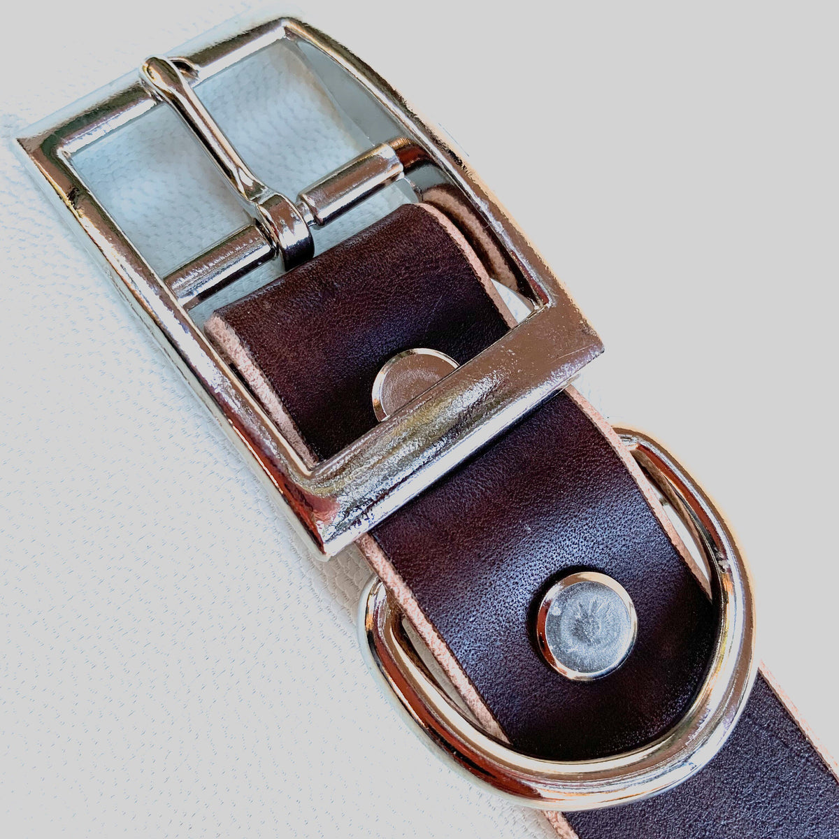 A close-up of a dark brown leather belt with a polished silver-tone metal buckle and keeper, set against a light textured background.
