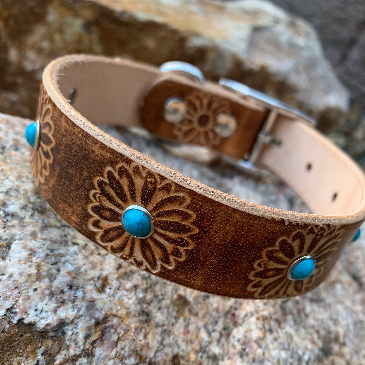 A brown leather dog collar with floral engravings and turquoise stone accents, resting on a textured rock with a blurred green garden background.