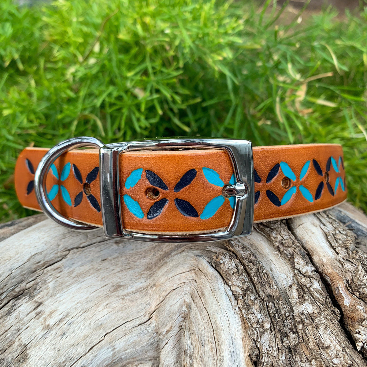 A tan leather dog collar with a silver buckle and D-ring, decorated with a hand-painted teal and navy blue geometric pattern, resting on a weathered log with green grass in the background.
