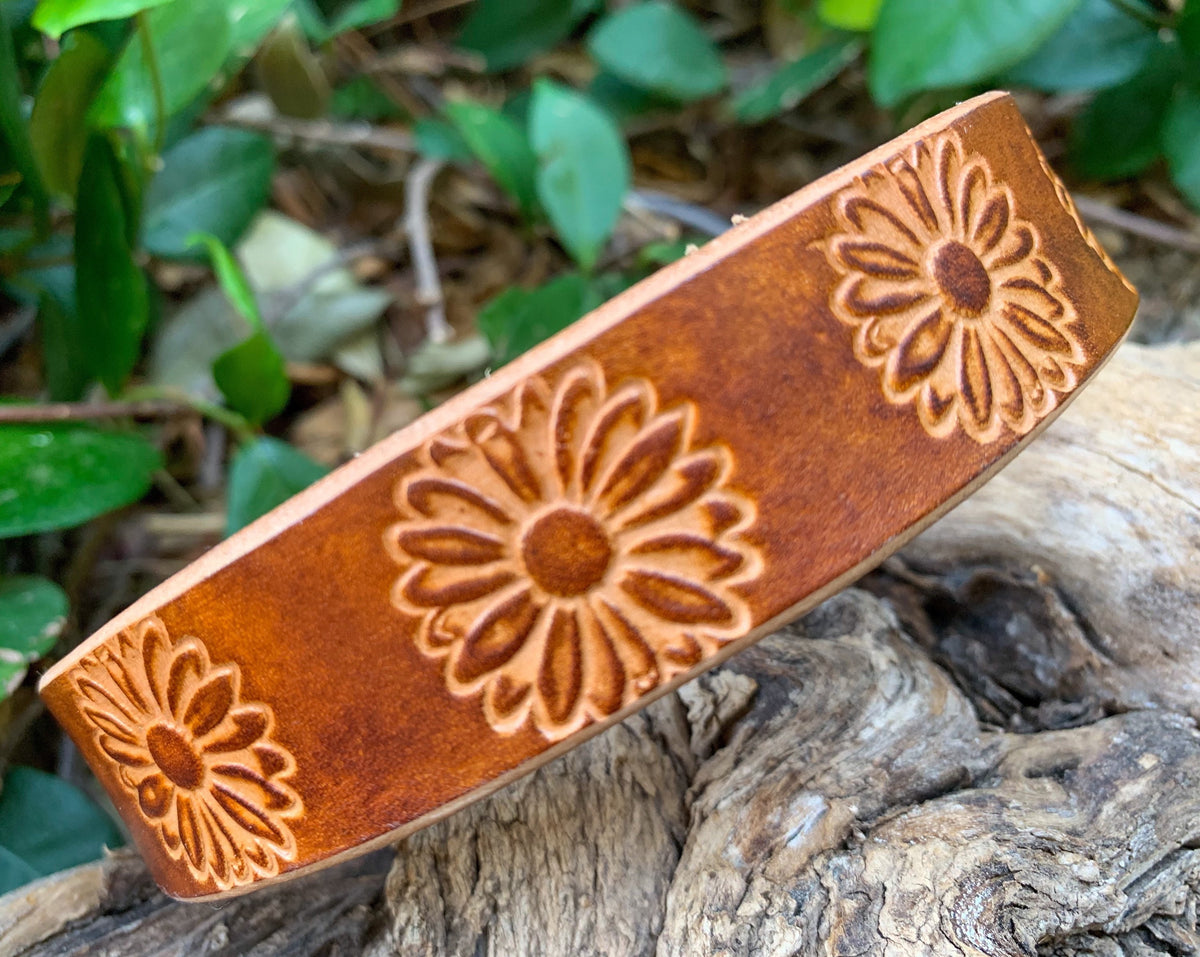 A brown leather dog collar with embossed daisy flower patterns, resting on a piece of weathered wood with green foliage in the background.