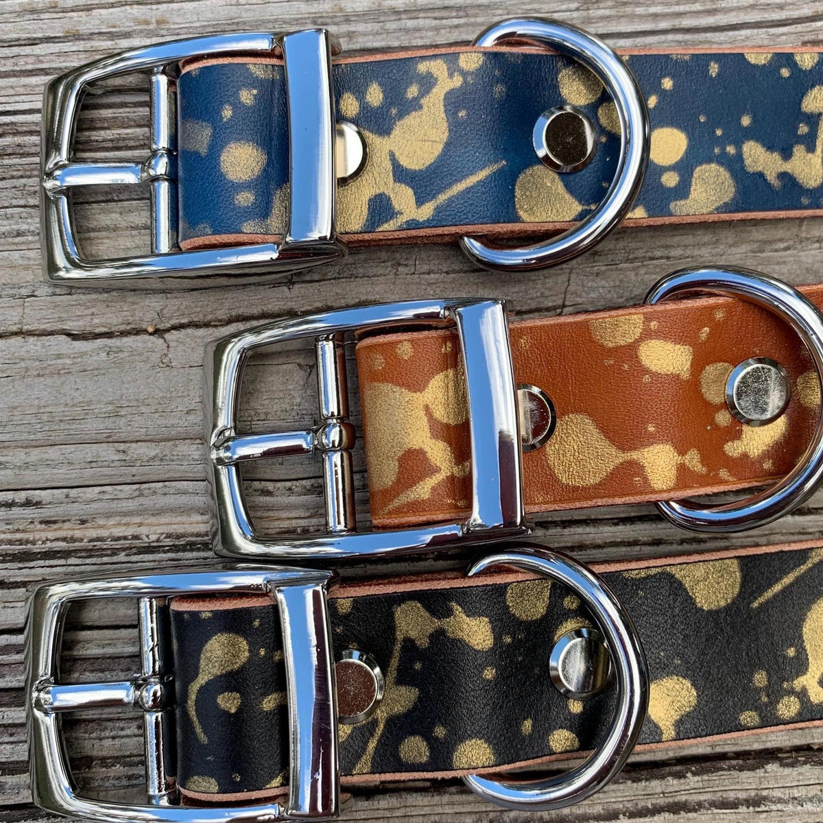 Three leather dog collars with polished silver-tone metal buckles and D-rings, arranged diagonally on a weathered wooden surface. The top collar is navy blue with gold paint splatter, the middle is tan/caramel with gold paint splatter, and the bottom is black with gold paint splatter.