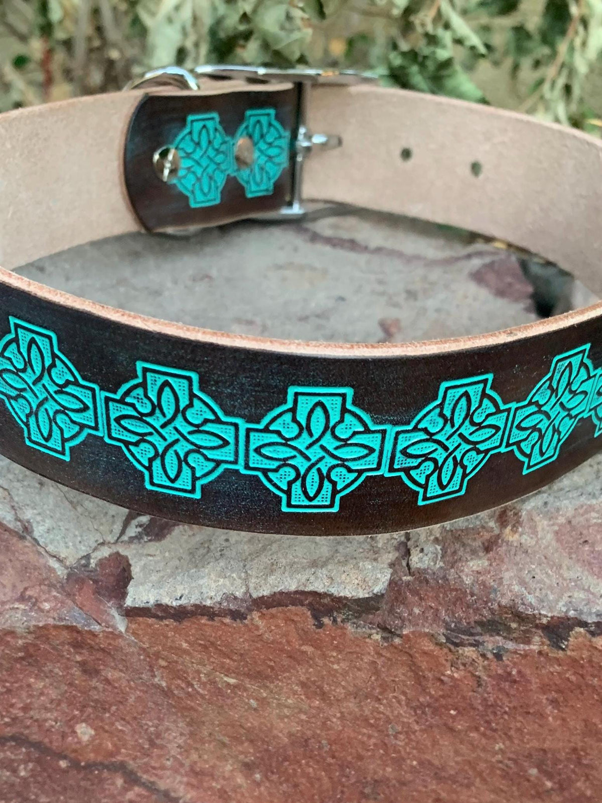 A dark brown leather collar with a repeating turquoise Celtic knot pattern, resting on a stone surface with a blurred natural background.